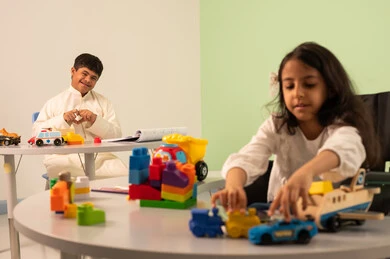 Creativity and development of mental and intellectual skills in Saudi kindergartens, Saudi society's interest in people of determination, recreational activities for people with special needs, a Saudi Arabian girl wearing casual clothes sitting in a wheelchair, enjoying games with happy expressions, encouraging, caring for and developing children's skills, an Omani boy with Down syndrome making a heart-shaped gesture with his hand