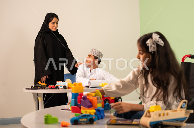 Expressions of happiness and pleasure, having a good time, developing mental and intellectual skills in Saudi kindergartens, a Saudi Arabian Gulf girl wearing casual clothes sitting in a wheelchair and playing with blocks, the concept of interest in hobbies, a Saudi boy with Down syndrome drawing and coloring with colors accompanied by his teacher