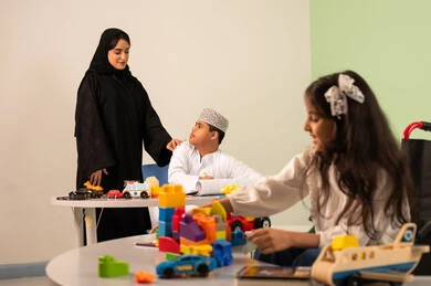 Expressions of happiness and pleasure, having a good time, developing mental and intellectual skills in Saudi kindergartens, a Saudi Arabian Gulf girl wearing casual clothes sitting in a wheelchair and playing with blocks, the concept of interest in hobbies, a Saudi boy with Down syndrome drawing and coloring with colors accompanied by his teacher