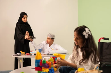 Gestures of concentration and enjoyment of drawing and coloring, having a good time, developing mental and intellectual skills in Saudi kindergartens, a Saudi Arabian Gulf girl wearing casual clothes sitting in a wheelchair and playing with blocks, the concept of interest in hobbies, a Saudi boy with Down syndrome drawing and coloring with colors accompanied by his teacher