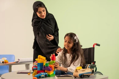 Having fun, developing mental and intellectual skills in Saudi kindergartens, expressions of happiness and pleasure, a Saudi Arabian Gulf girl wearing casual clothes sitting in a wheelchair and playing with blocks, the concept of interest in hobbies