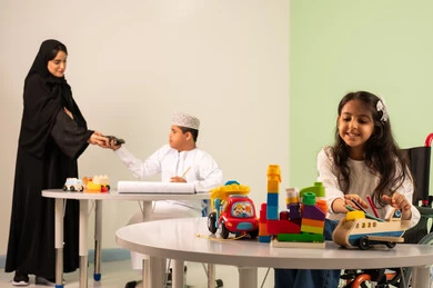 Developing mental and intellectual skills in Saudi kindergartens, gestures of concentration and enjoyment of drawing and coloring, spending enjoyable times, a Saudi Arabian Gulf girl wearing casual clothes sitting in a wheelchair and playing with blocks, the concept of interest in hobbies, a Saudi boy with Down syndrome drawing and coloring with colors accompanied by his teacher