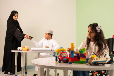 Gestures of concentration and enjoyment of drawing and coloring, having a good time, developing artistic skills in Saudi kindergartens, a Saudi Arabian Gulf girl wearing casual clothes sitting in a wheelchair and playing with blocks, the concept of interest in hobbies, a Saudi boy with Down syndrome drawing and coloring with colors accompanied by his teacher