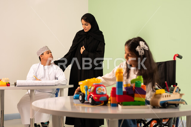 Having fun, developing artistic skills in Saudi kindergartens, a Saudi Arabian Gulf girl wearing casual clothes sitting on a wheelchair playing with blocks, the concept of interest in hobbies, gestures of concentration and enjoyment of drawing and coloring, a Saudi boy with Down syndrome drawing and coloring with colors accompanied by his teacher