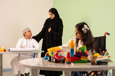Having fun, developing artistic skills in Saudi kindergartens, a Saudi Arabian Gulf girl wearing casual clothes sitting on a wheelchair playing with blocks, the concept of interest in hobbies, gestures of concentration and enjoyment of drawing and coloring, a Saudi boy with Down syndrome drawing and coloring with colors accompanied by his teacher