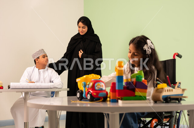 تنمية المهارات الفنية في الروضات السعودية ، بنت عربية خليجية سعودية ترتدي الزي الكاجوال تجلس على كرسي متحرك وتقوم باللعب بالمكعبات ،مفهوم الاهتمام بالهوايات، إيماءات التركيز والاستمتاع بالرسم و التلوين،ولد سعودي مصاب بمتلازمه داون يقوم بالرسم والتلوين بالألوان برفقه معلمته، قضاء اوقات ممتعه