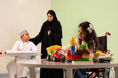 Developing artistic skills in Saudi kindergartens, a Saudi Arabian Gulf girl wearing casual clothes sitting on a wheelchair playing with blocks, the concept of interest in hobbies, gestures of concentration and enjoyment of drawing and coloring, a Saudi boy with Down syndrome drawing and coloring with colors accompanied by his teacher, spending enjoyable times