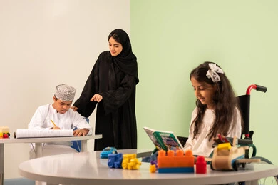The concept of interest in hobbies, a Saudi Arabian Gulf girl wearing casual clothes sitting on a wheelchair playing with blocks, behind her a Saudi boy with Down syndrome is drawing and coloring with colors accompanied by his teacher, gestures of concentration and enjoyment of drawing and coloring, spending fun times, developing artistic skills in Saudi kindergartens