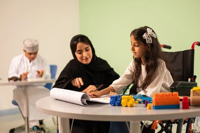 Gestures of concentration and enjoyment of drawing and coloring, having a good time, developing artistic skills in Saudi kindergartens, a Saudi Arabian Gulf girl wearing casual clothes sitting on a wheelchair and drawing and coloring with colors accompanied by her teacher, behind them a Saudi boy with Down syndrome, the concept of interest in hobbies