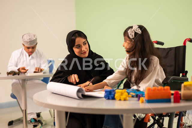 قضاء اوقات ممتعه ، تنمية المهارات الفنية في الروضات السعودية ، بنت عربية خليجية سعودية ترتدي الزي الكاجوال تجلس على كرسي متحرك وتقوم بالرسم والتلوين بالألوان برفقه معلمتها ،بخلفهم ولد سعودي مصاب بمتلازمه داون ، مفهوم الاهتمام بالهوايات,  إيماءات التركيز والاستمتاع بالرسم و التلوين