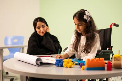 تنمية المهارات الفنية في الروضات السعودية ، بنت عربية خليجية سعودية ترتدي الزي الكاجوال تجلس على كرسي متحرك وتقوم بالرسم والتلوين بالألوان برفقه معلمتها ، قضاء اوقات ممتعه ، مفهوم الاهتمام بالهوايات,  إيماءات التركيز والاستمتاع بالرسم و التلوين