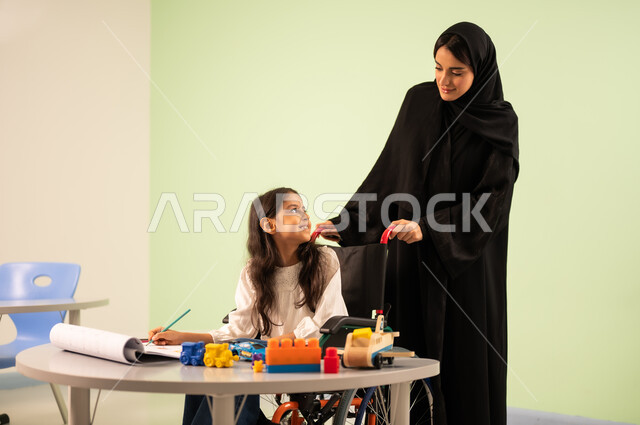 مفهوم الاهتمام بالهوايات, تنمية المهارات الفنية في الروضات السعودية ،  إيماءات التركيز والاستمتاع بالرسم و التلوين،بنت عربية خليجية سعودية ترتدي الزي الكاجوال تجلس على كرسي متحرك وتقوم بالرسم والتلوين بالألوان برفقه معلمتها ، قضاء اوقات ممتعه