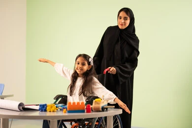 تنمية المهارات الفنية في الروضات السعودية ،  إيماءات التركيز والاستمتاع بالرسم و التلوين،مفهوم الاهتمام بالهوايات, بنت عربية خليجية سعودية ترتدي الزي الكاجوال تجلس على كرسي متحرك وتقوم بالرسم والتلوين بالألوان برفقه معلمتها ، قضاء اوقات ممتعه