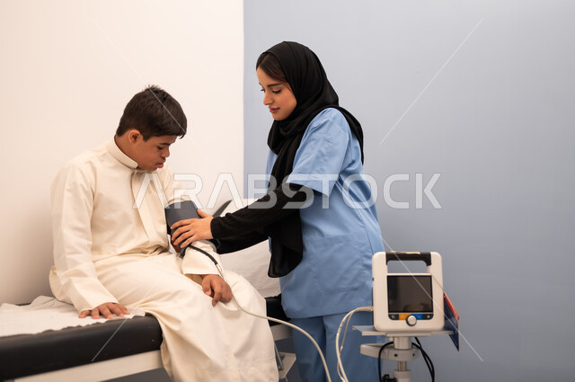 المعاناه من مشاكل صحية ، المتابعة الدورية لفحص الدم, تقديم أفضل الخدمات الصحية لمرضى ذوى الاحتياجات الخاصه ، مفهوم الطب والرعاية الصحية، ولد عربي سعودي خليجي مصاب بمتلازمه داون يجلس على كرسي العلاج فى المستشفي ، ممرضه عمانيه تقوم بقياس ضغط الدم باستخدام الميزان الالكتروني