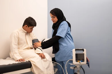 المعاناه من مشاكل صحية ، المتابعة الدورية لفحص الدم, تقديم أفضل الخدمات الصحية لمرضى ذوى الاحتياجات الخاصه ، مفهوم الطب والرعاية الصحية، ولد عربي سعودي خليجي مصاب بمتلازمه داون يجلس على كرسي العلاج فى المستشفي ، ممرضه عمانيه تقوم بقياس ضغط الدم باستخدام الميزان الالكتروني