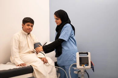 المتابعة الدورية لفحص الدم, تقديم أفضل الخدمات الصحية لمرضى ذوى الاحتياجات الخاصه ، مفهوم الطب والرعاية الصحية، ولد عربي سعودي خليجي مصاب بمتلازمه داون يجلس على كرسي العلاج فى المستشفي ، ممرضه عمانيه تقوم بقياس ضغط الدم باستخدام الميزان الالكتروني،المعاناه من مشاكل صحية