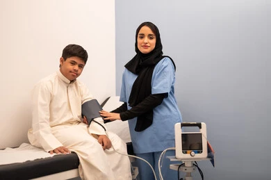 مفهوم الطب والرعاية الصحية، المتابعة الدورية لفحص الدم, تقديم أفضل الخدمات الصحية لمرضى ذوى الاحتياجات الخاصه ، ولد عربي سعودي خليجي مصاب بمتلازمه داون يجلس على كرسي العلاج فى المستشفي ، ممرضه عمانيه تقوم بقياس ضغط الدم باستخدام الميزان الالكتروني،المعاناه من مشاكل صحية