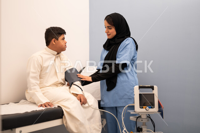 تقديم أفضل الخدمات الصحية لمرضى ذوى الاحتياجات الخاصه ، مفهوم الطب والرعاية الصحية، المتابعة الدورية لفحص الدم, ولد عربي سعودي خليجي مصاب بمتلازمه داون يجلس على كرسي العلاج فى المستشفي ، ممرضه عمانيه تقوم بقياس ضغط الدم باستخدام الميزان الالكتروني،المعاناه من مشاكل صحية