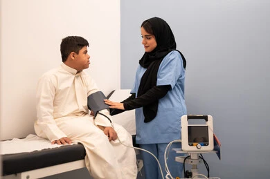 تقديم أفضل الخدمات الصحية لمرضى ذوى الاحتياجات الخاصه ، مفهوم الطب والرعاية الصحية، المتابعة الدورية لفحص الدم, ولد عربي سعودي خليجي مصاب بمتلازمه داون يجلس على كرسي العلاج فى المستشفي ، ممرضه عمانيه تقوم بقياس ضغط الدم باستخدام الميزان الالكتروني،المعاناه من مشاكل صحية