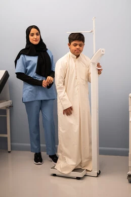 مفهوم الاهتمام بالصحه واللياقه،الوزن الزائد والسمنة ، تراكم السعرات الحرارية ، ولد عربي سعودي خليجي مصاب بمتلازمه داون ممتلئ الجسم يعاني من السمنة ، ممرضه عمانيه تستخدم ميزان لقياس الوزن ،المعاناه من مشاكل صحية