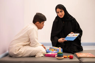 وسائل ايضاح ترفيفهيه للتعليم ،ايماءات التركيز والاندماج  ،تنمية المهارات الذهنيه والفكريه في الروضات السعودية ،ولد عربي سعودي مصاب بمتلازمه داون يقوم باللعب بلعبه الكروت التعليميه برفقه معلمته ، مفهوم التعلم عن طريق اللعب، قضاء اوقات ممتعه