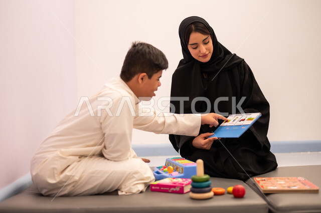 Developing mental and intellectual skills in Saudi kindergartens, a Saudi Arabian boy with Down syndrome playing an educational card game with his teacher, the concept of learning through play, having fun, educational entertainment means, expressions of pleasure and happiness, gestures of concentration and integration
