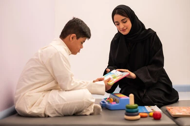 قضاء اوقات ممتعه  ، تنمية المهارات الذهنيه والفكريه في الروضات السعودية ،وسائل ايضاح ترفيفهيه للتعليم ،تعابير السرور والسعاده ، ايماءات التركيز والاندماج ، تعزيز النشاط الذهني، مفهوم هوايه المطالعه ، ولد عربي سعودي مصاب بمتلازمه داون يقوم باللعب بلعبه الكروت التعليميه برفقه معلمته