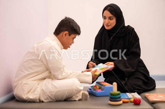 وسائل ايضاح ترفيفهيه للتعليم ،تعابير السرور والسعاده ، ايماءات التركيز والاندماج ، تعزيز النشاط الذهني، مفهوم هوايه المطالعه ، ولد عربي سعودي مصاب بمتلازمه داون يقوم باللعب بلعبه الكروت التعليميه برفقه معلمته، تنمية المهارات الذهنيه والفكريه في الروضات السعودية ،قضاء اوقات ممتعه