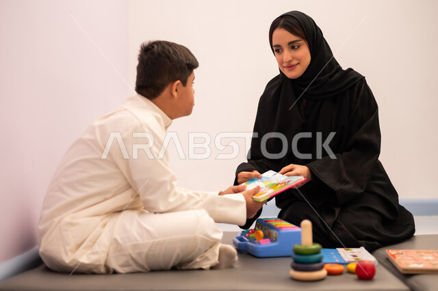 Gestures of concentration and integration, enhancing mental activity, the concept of the hobby of reading, a Saudi Arabian boy with Down syndrome playing an educational card game with his teacher, developing mental and intellectual skills in Saudi kindergartens, educational entertainment means, expressions of pleasure and happiness, spending a pleasant time