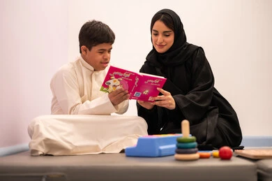Enhancing mental activity, gestures of concentration and integration, the concept of the hobby of reading, a Saudi Arabian boy with Down syndrome playing an educational card game with his teacher, developing mental and intellectual skills in Saudi kindergartens, educational entertainment means, expressions of pleasure and happiness, spending enjoyable times