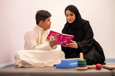 The concept of reading hobby, a Saudi Arabian boy with Down syndrome playing educational cards with his teacher, developing mental and intellectual skills in Saudi kindergartens, educational entertainment means, enhancing mental activity, gestures of concentration and integration, expressions of pleasure and happiness, spending enjoyable times