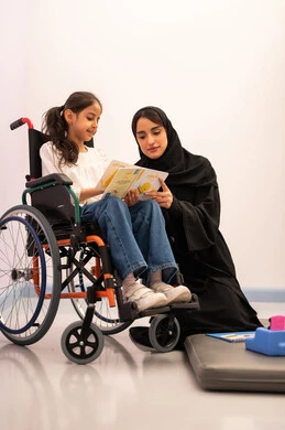 مفهوم هوايه المطالعه ،تعزيز النشاط الذهني، قضاء اوقات ممتعه ،تنمية المهارات الذهنيه والفكريه في الروضات السعودية ، بنت عربيه سعوديه ترتدي الزي الكاجوال تجلس على كرسي متحرك، تقوم بقراءه الكتب التعليميه الملونه برفقه معلمتها ، تعابير السرور والسعاده