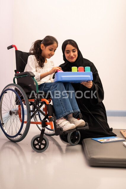 ايماءات التركيز والاندماج ، تنمية المهارات الذهنيه والفكريه في الروضات السعودية ، تعزيز النشاط الذهني، قضاء اوقات ممتعه ،بنت عربيه سعوديه ترتدي الزي الكاجوال تجلس على كرسي متحرك، تقوم باللعب بلعبه الكروت المطابقه برفقه معلمتها ، تعابير السرور والسعاده