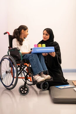 Developing mental and intellectual skills in Saudi kindergartens, enhancing mental activity, expressions of pleasure and happiness, spending enjoyable times, a Saudi Arabian girl wearing casual clothes sitting in a wheelchair, playing matching cards with her teacher, gestures of concentration and integration