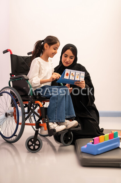 Expressions of pleasure and happiness, having a good time, a Saudi Arabian girl wearing casual clothes sitting on a wheelchair, playing matching cards with her teacher, gestures of concentration and integration, developing mental and intellectual skills in Saudi kindergartens, enhancing mental activity