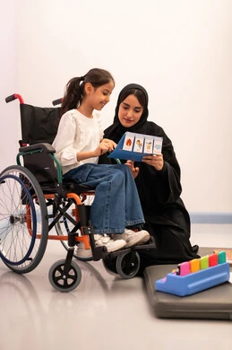 تعابير السرور والسعاده ، قضاء اوقات ممتعه ،بنت عربيه سعوديه ترتدي الزي الكاجوال تجلس على كرسي متحرك، تقوم باللعب بلعبه الكروت المطابقه برفقه معلمتها ، ايماءات التركيز والاندماج ، تنمية المهارات الذهنيه والفكريه في الروضات السعودية ، تعزيز النشاط الذهني