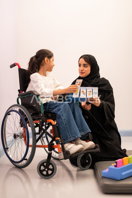 قضاء اوقات ممتعه ،تعزيز النشاط الذهني، ايماءات التركيز والاندماج ، تنمية المهارات الذهنيه والفكريه في الروضات السعودية ،  تعابير السرور والسعاده ، بنت عربيه سعوديه ترتدي الزي الكاجوال تجلس على كرسي متحرك، تقوم باللعب بلعبه الكروت المطابقه برفقه معلمتها