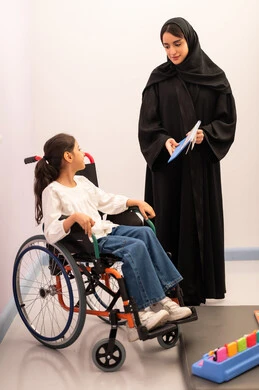 تعزيز النشاط الذهني، ايماءات التركيز والاندماج ، تنمية المهارات الذهنيه والفكريه في الروضات السعودية ،  تعابير السرور والسعاده ، قضاء اوقات ممتعه ،بنت عربيه سعوديه ترتدي الزي الكاجوال تجلس على كرسي متحرك، تقوم باللعب بلعبه الكروت المطابقه برفقه معلمتها