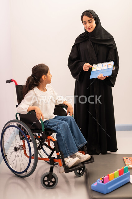 Gestures of concentration and integration, developing mental and intellectual skills in Saudi kindergartens, enhancing mental activity, expressions of pleasure and happiness, spending enjoyable times, a Saudi Arabian girl wearing casual clothes sitting in a wheelchair, playing the matching card game with her teacher