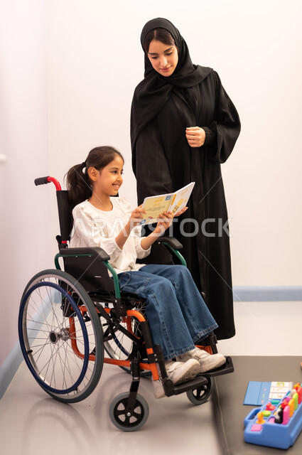 Developing mental and intellectual skills in Saudi kindergartens, having fun, enhancing mental activity, the concept of reading hobby, a Saudi Arabian girl wearing casual clothes sitting in a wheelchair, reading colorful educational books with her teacher, expressions of pleasure and happiness