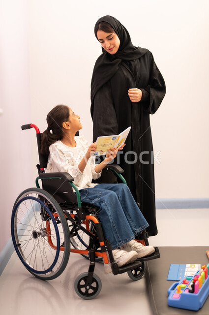Having fun, developing mental and intellectual skills in Saudi kindergartens, enhancing mental activity, the concept of reading hobby, a Saudi Arabian girl wearing casual clothes sitting on a wheelchair, reading colorful educational books with her teacher, expressions of pleasure and happiness