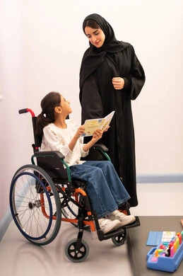 قضاء اوقات ممتعه ، تنمية المهارات الذهنيه والفكريه في الروضات السعودية ، تعزيز النشاط الذهني،  مفهوم هوايه المطالعه ،بنت عربيه سعوديه ترتدي الزي الكاجوال تجلس على كرسي متحرك، تقوم بقراءه الكتب التعليميه الملونه برفقه معلمتها ، تعابير السرور والسعاده