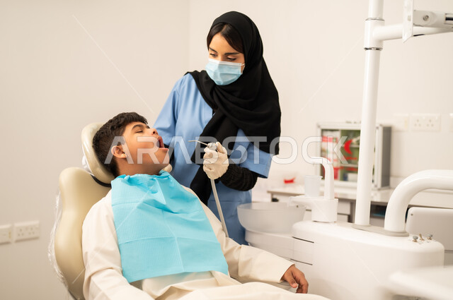 The concept of medicine and healthcare, using sterile instruments for dental examinations, providing medical services to patients, oral health care, a Saudi boy with Down syndrome sitting on a treatment chair, a female doctor wearing gloves and a mask examining the patient's teeth.