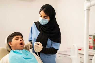 مفهوم الطب والرعاية الصحية، طبيبة ترتدي القفازات والكمامه تقوم بفحص أسنان المريض، تقديم الخدمات الطبية للمرضى,ولد سعودي مصاب بمتلازمة داون  يجلس على كرسي العلاج ، استخدام الأدوات المعقمة لإجراء الفحوصات السنية،العناية بالصحة الفموية