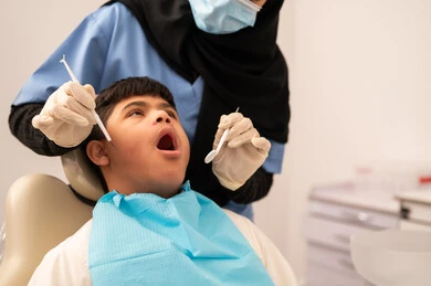 تقديم الخدمات الطبية للمرضى, ولد سعودي مصاب بمتلازمة داون  يجلس على كرسي العلاج ، استخدام الأدوات المعقمة لإجراء الفحوصات السنية،العناية بالصحة الفموية، طبيبة ترتدي القفازات والكمامه تقوم بفحص أسنان المريض، مفهوم الطب والرعاية الصحية