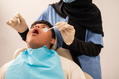 طبيبة ترتدي القفازات والكمامه تقوم بفحص أسنان المريض، تقديم الخدمات الطبية للمرضى,ولد سعودي مصاب بمتلازمة داون  يجلس على كرسي العلاج ، استخدام الأدوات المعقمة لإجراء الفحوصات السنية،العناية بالصحة الفموية،مفهوم الطب والرعاية الصحية