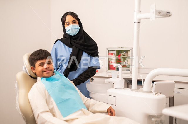The concept of medicine and healthcare, using sterile instruments for dental examinations, oral health care, a female doctor wearing gloves and a mask examining a patient's teeth, providing medical services to patients, a Saudi boy with Down syndrome sitting on a treatment chair looking at the camera with a joyful expression.
