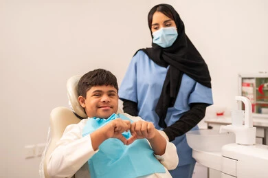 تقديم الخدمات الطبية للمرضى,  ولد سعودي مصاب بمتلازمة داون  يجلس على كرسي العلاج يشير بيده على شكل قلب، طبيبة ترتدي القفازات والكمامه تقوم بفحص أسنان المريض، مفهوم الطب والرعاية الصحية، استخدام الأدوات المعقمة لإجراء الفحوصات السنية،العناية بالصحة الفموية