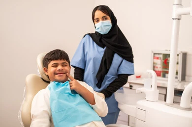 ولد سعودي مصاب بمتلازمة داون  يجلس على كرسي العلاج ينظر الى الكاميرا بتعابير البهجه ، طبيبة ترتدي القفازات والكمامه تقوم بفحص أسنان المريض، تقديم الخدمات الطبية للمرضى,  مفهوم الطب والرعاية الصحية، استخدام الأدوات المعقمة لإجراء الفحوصات السنية،العناية بالصحة الفموية