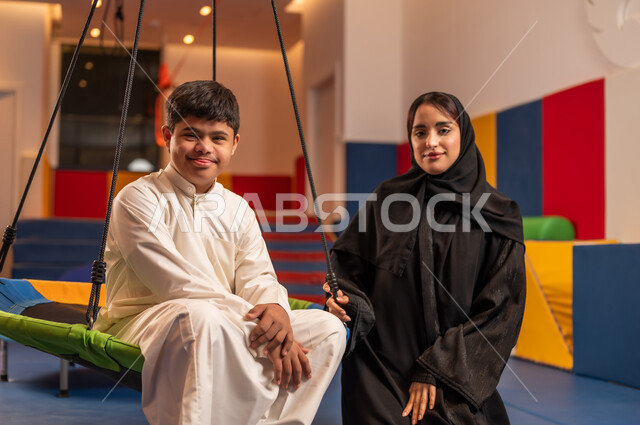 Enjoying summer vacation, times of entertainment and fun, gestures of pleasure and happiness, having a wonderful time at an amusement park, a Saudi boy with Down syndrome at an amusement park with his mother, playing on the swings.